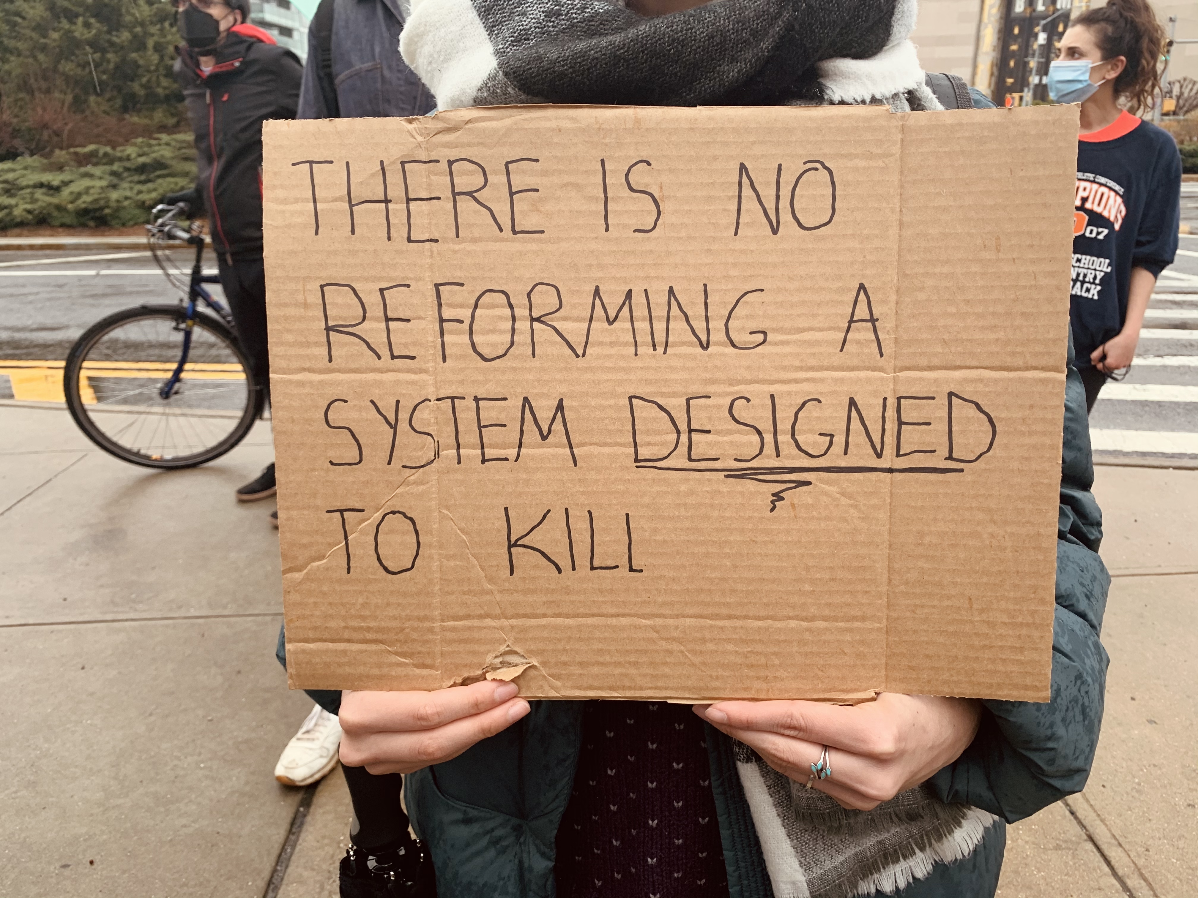 Fotografía en color tomada en la calle durante una manifestación. En el centro de la imagen, una persona sostiene ante sí una pancarta de cartón marrón que le cubre el rostro y el torso. Sobre el cartón, escrito a mano con rotulador negro en letras mayúsculas y en inglés, se lee: «No se puede reformar un sistema diseñado para matar», con la palabra «diseñado» subrayada. Solo se le ven las manos, que sujetan el cartón por los lados, y las piernas. Al fondo, desenfocadas, hay otras personas con mascarilla, una bicicleta apoyada y una acera con árboles.