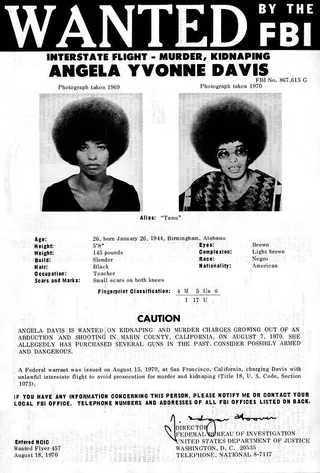 Cartel de búsqueda del FBI de 1970. En la cabecera, en letras grandes, «WANTED BY THE FBI», y debajo los cargos: fuga interestatal, asesinato y secuestro. A continuación el nombre, Angela Yvonne Davis, y su número de expediente. En el centro, dos fotografías de una mujer joven negra con peinado afro, tomadas en 1969 y 1970. Bajo ellas, una tabla con la descripción física: edad, fecha y lugar de nacimiento, estatura, peso, complexión, pelo, ojos, ocupación (maestra) y una casilla de raza que usa el término «Negro», habitual en los documentos oficiales estadounidenses de la época. Sigue un párrafo de advertencia que la describe como posiblemente armada y peligrosa, y al pie la firma del director del FBI y la fecha, 18 de agosto de 1970.