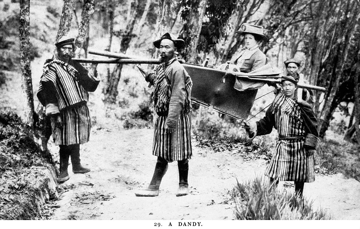 Postal en blanco y negro: una mujer europea sentada en una hamaca de lona sujeta a un bastidor, llevada a hombros por porteadores indios.
