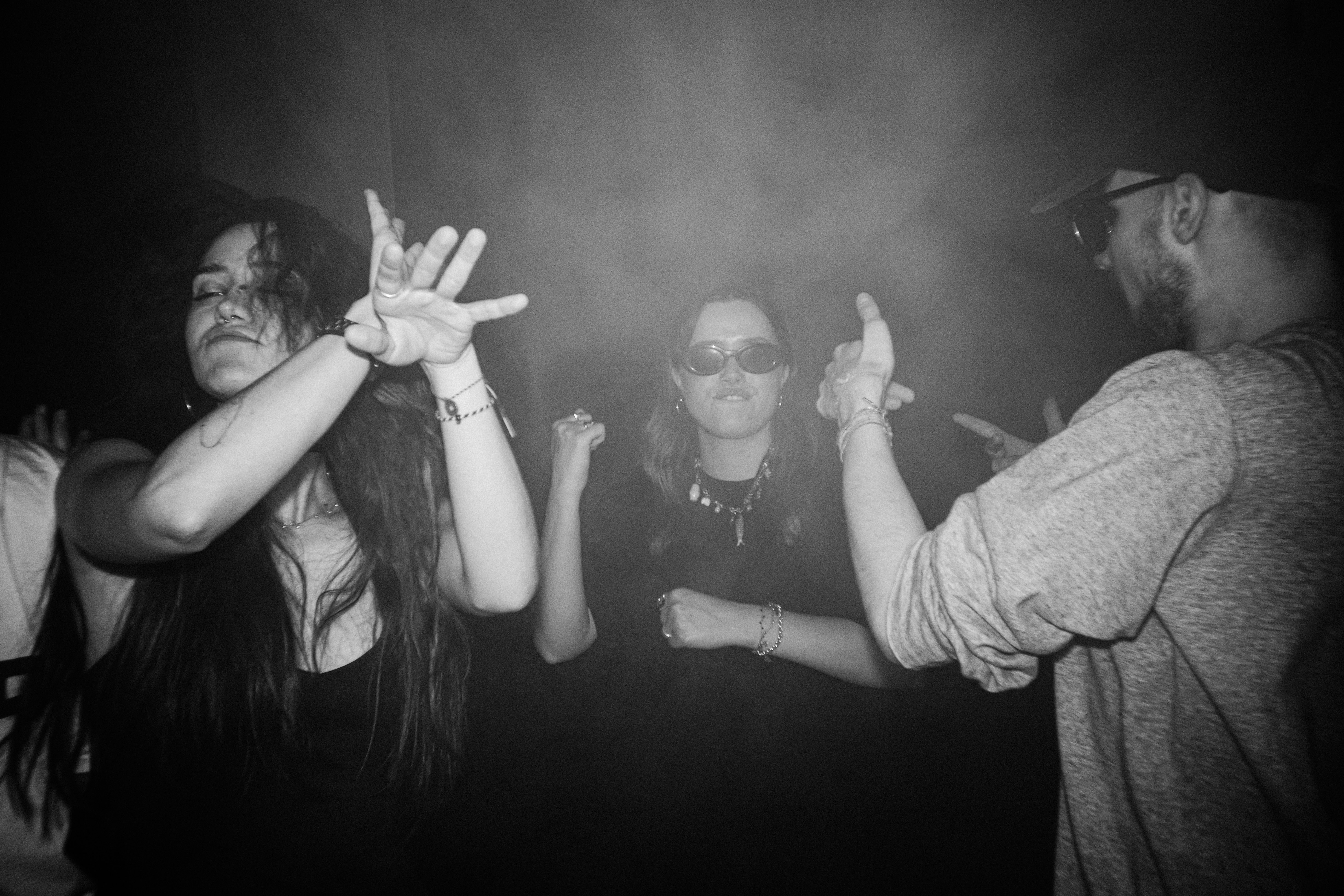 Interior de una sala en penumbra, con humo y luces de colores, y varias personas bailando.